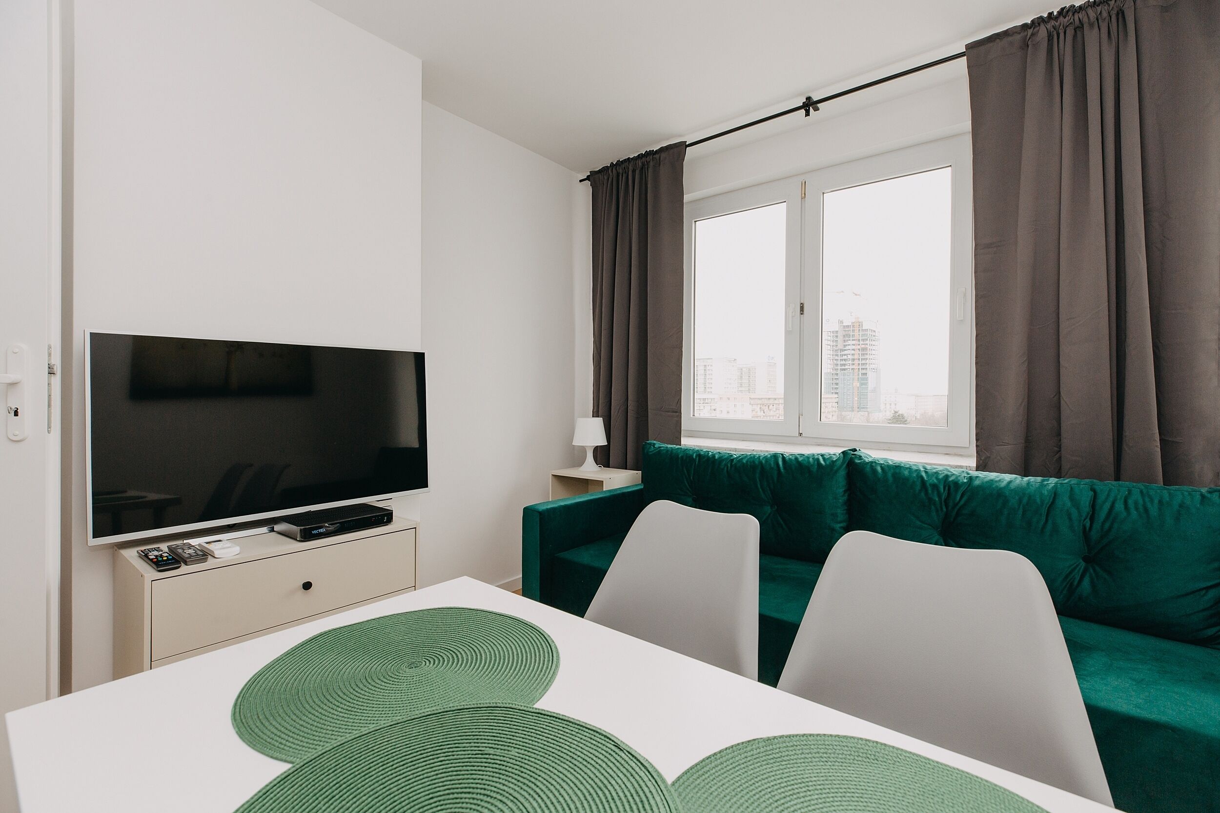 A modern hotel room with a flat-screen TV, a green sofa, a white dining table, and a view of the city through large windows.