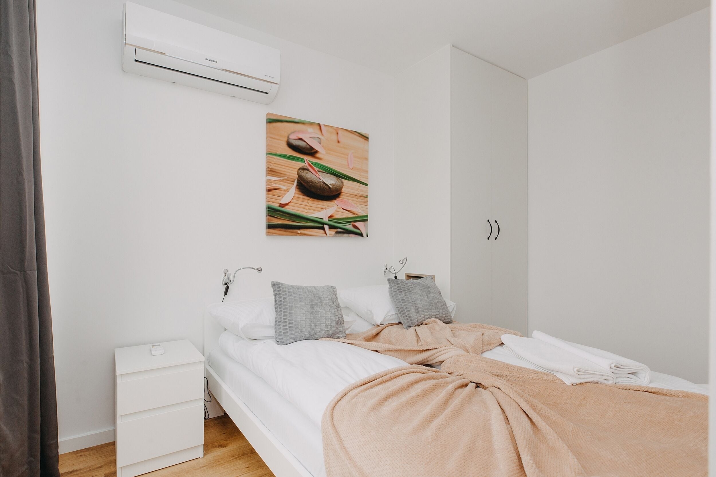 A bedroom with a bed, white nightstand, and a wall-mounted air conditioner.