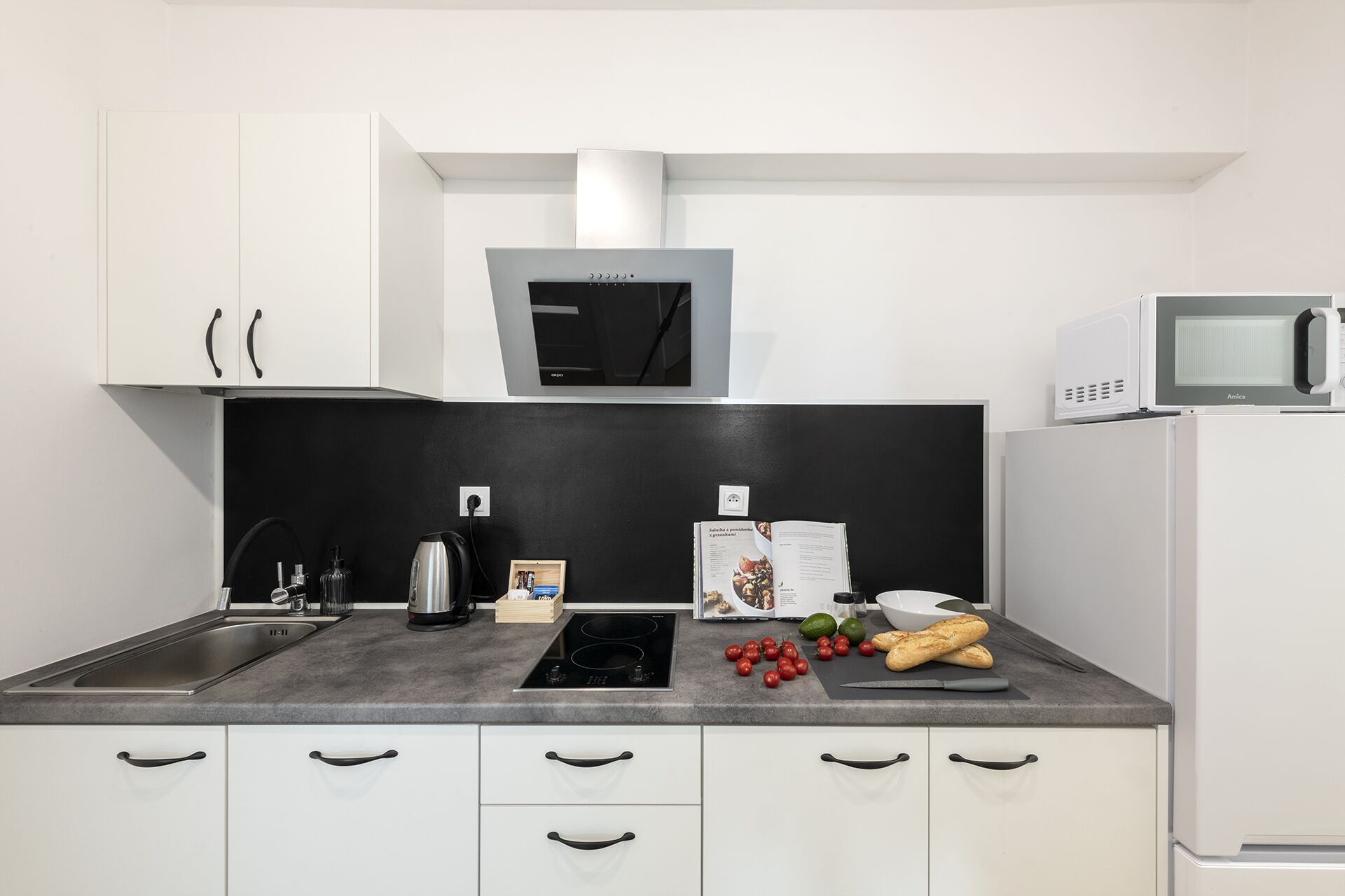 A modern kitchen with white cabinets, a black backsplash, a built-in oven, a microwave, and a countertop with a kettle, a cutting board, tomatoes, and bread.