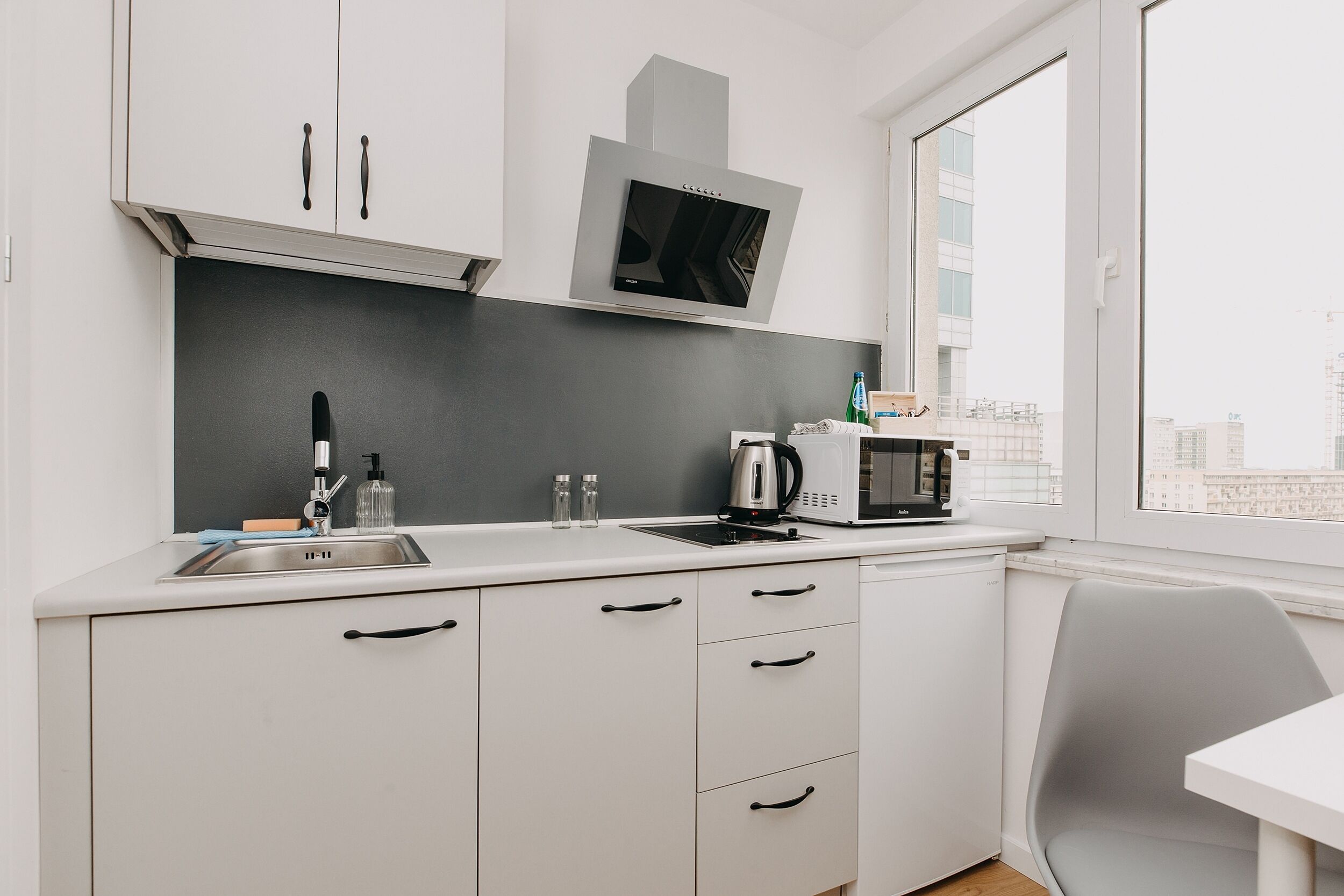 A modern kitchen with white cabinets, a microwave, a kettle, and a window with a city view.