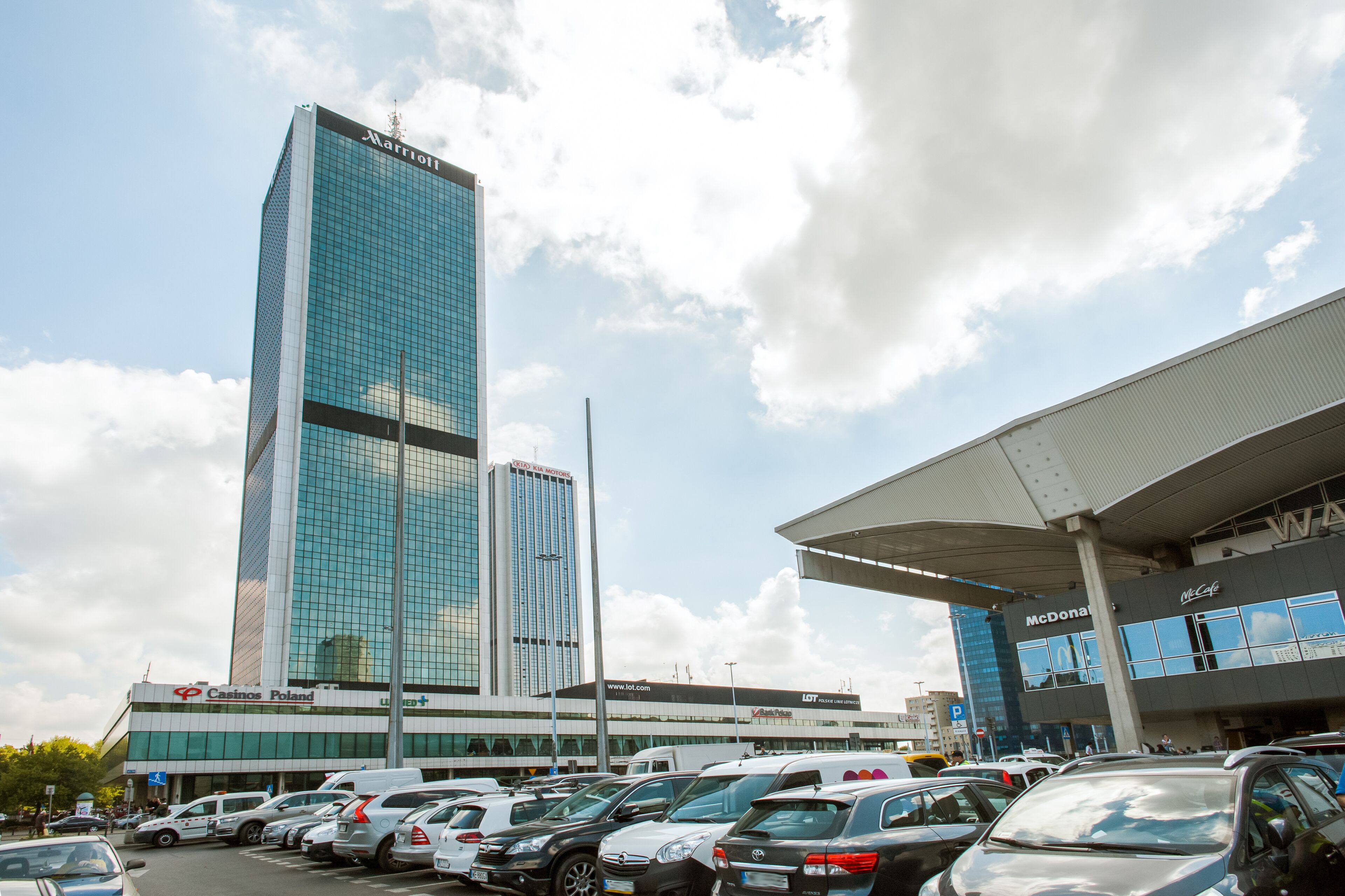Een hotel met een moderne glazen gevel, een parkeerplaats met diverse voertuigen en een heldere lucht.