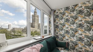 A room with a large window overlooking a cityscape, a green sofa, and tropical wallpaper. - ShortStayPoland Panska (Warsaw)