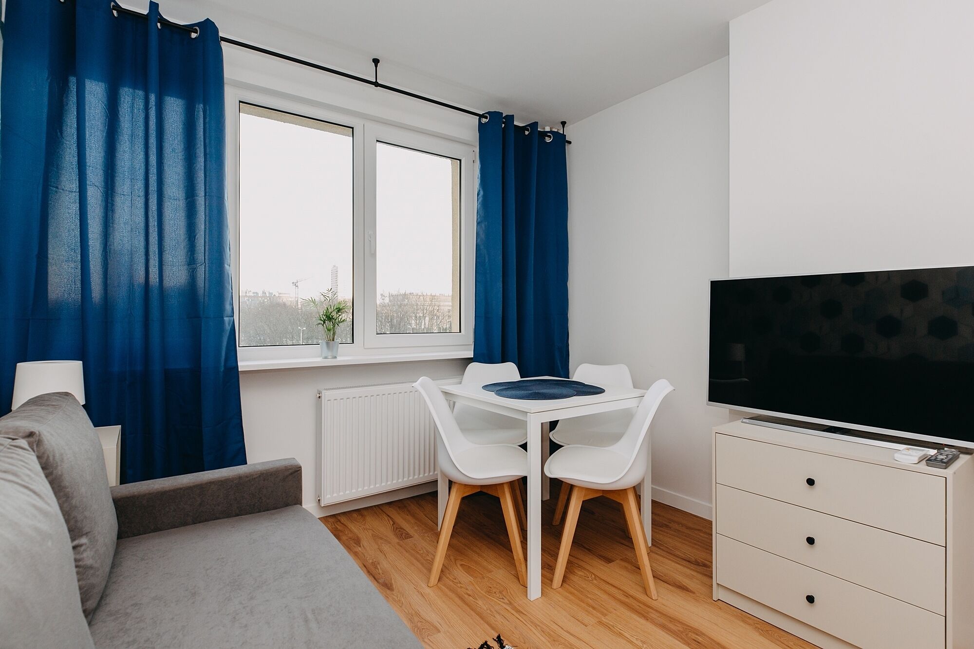 Una habitación moderna y bien iluminada con un sofá gris, una mesa blanca con dos sillas, un televisor sobre un mueble y una ventana con cortinas azules.