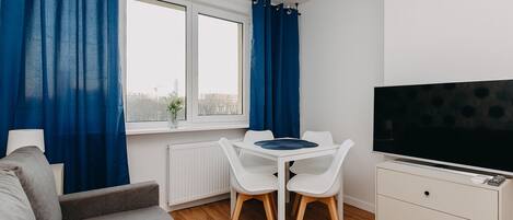 A modern, well-lit room with a grey sofa, a white table with two chairs, a TV on a cabinet, and a window with blue curtains.