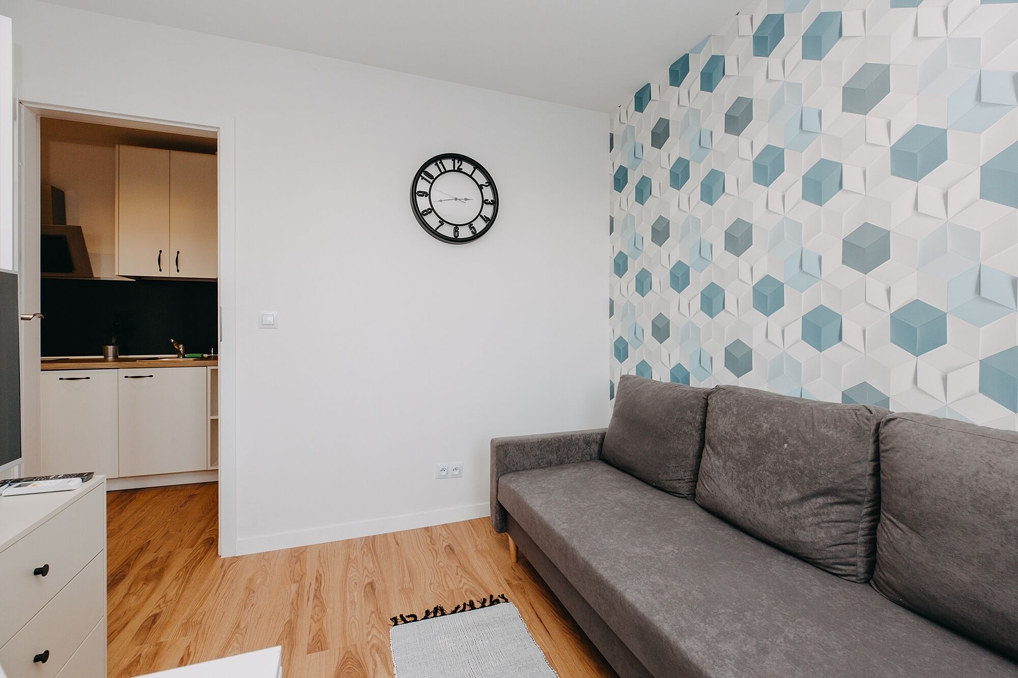 Una sala de estar moderna con un sofá gris, un pared con estampado geométrico y una cocina minimalista visible a través de una puerta abierta.