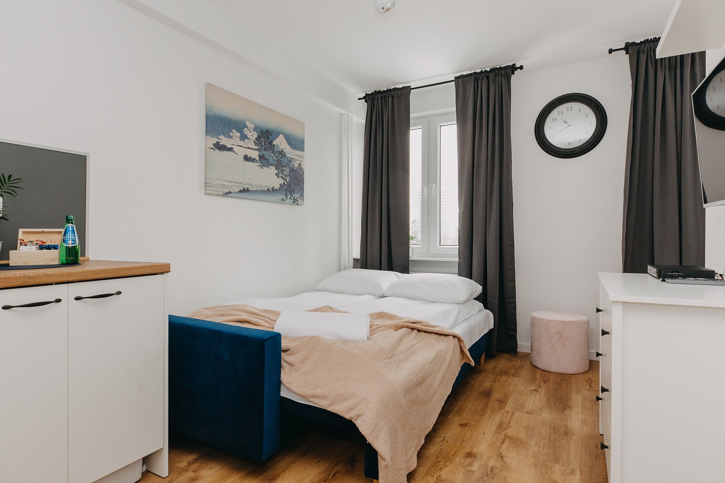 Un dormitorio con cama, escritorio, silla, taburete pequeño, un reloj de pared y un cuadro.