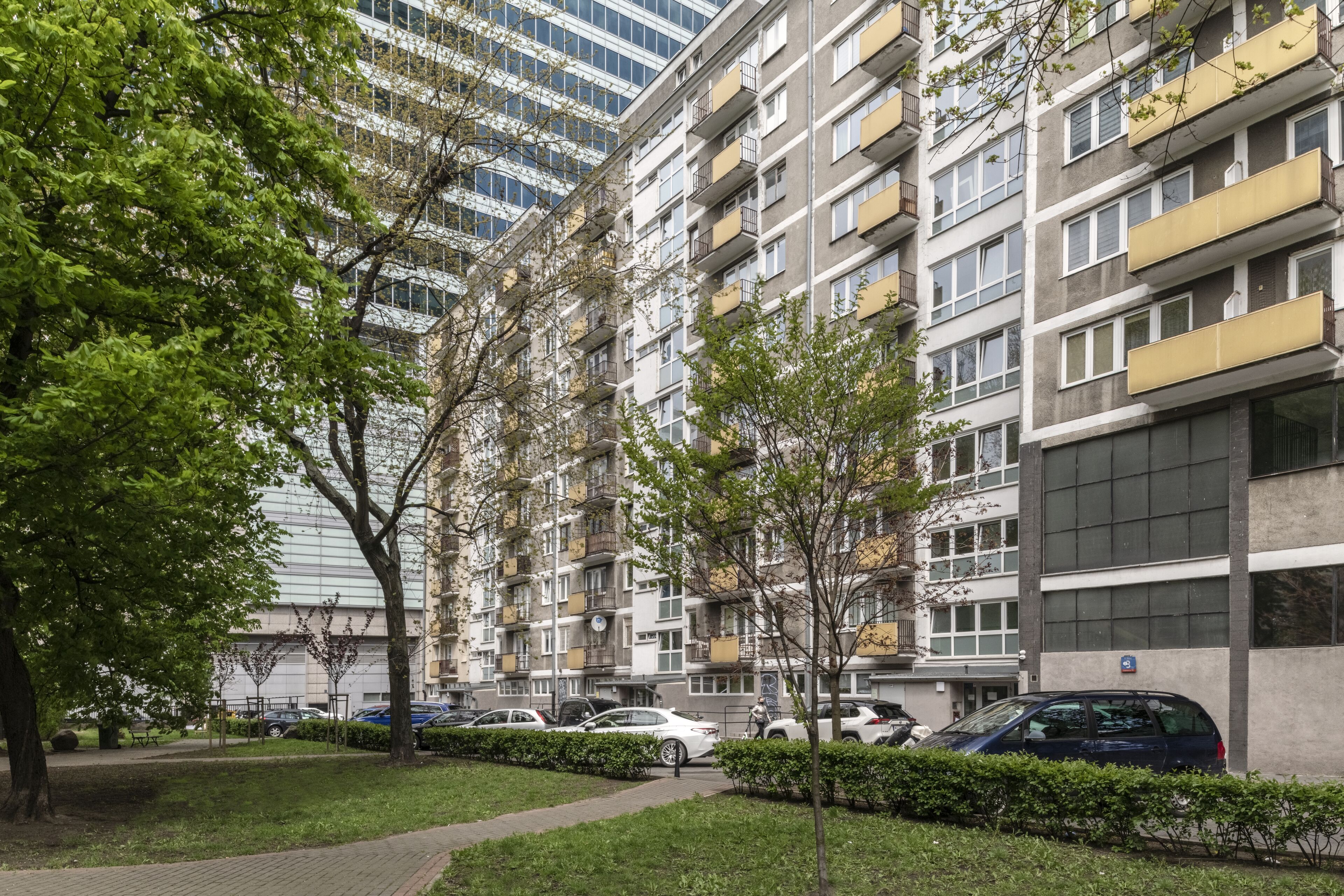 Un edificio residencial de varios pisos con balcones, un sendero pavimentado y automóviles estacionados.