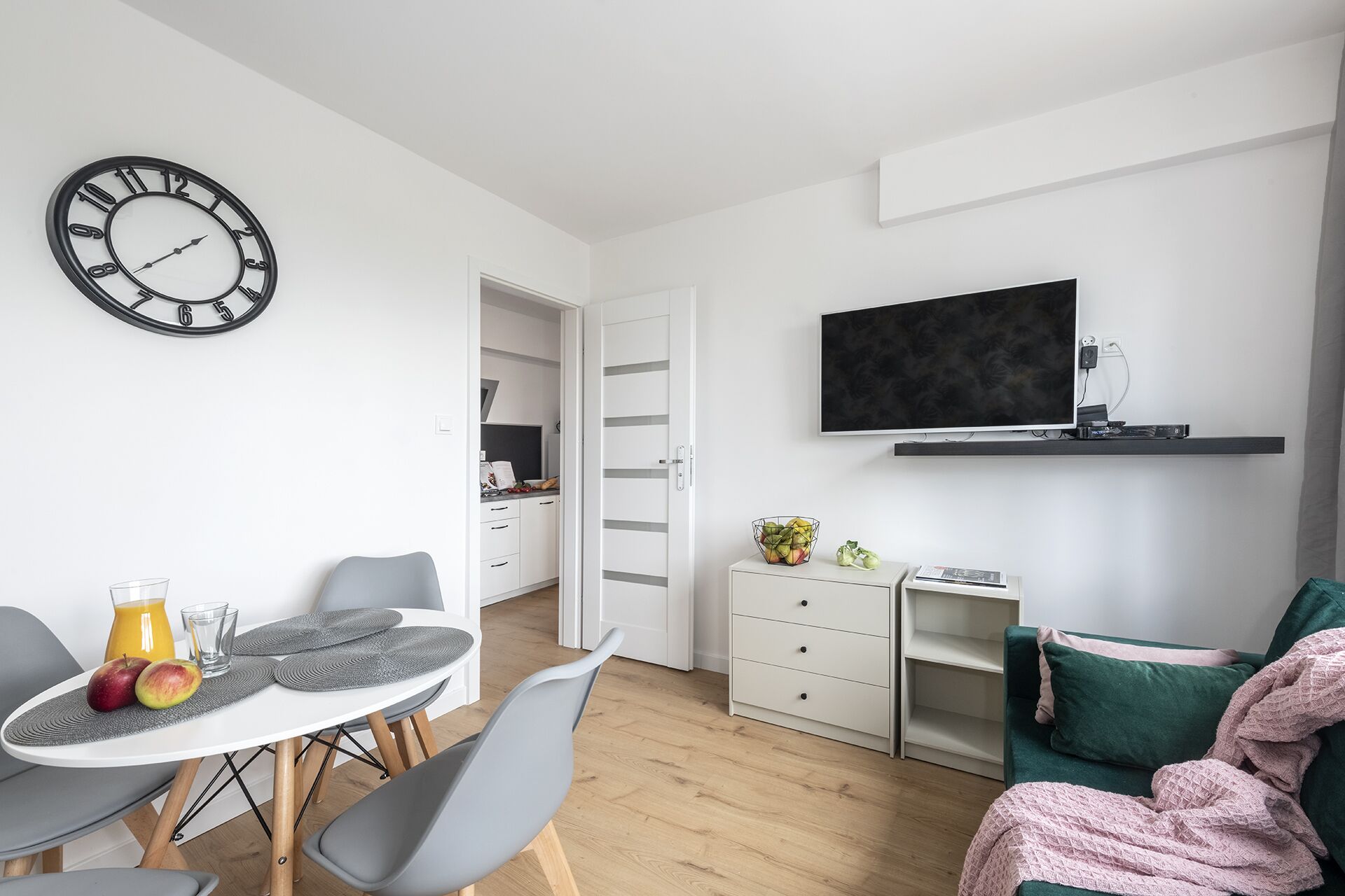 A modern living room with a dining area, a TV mounted on the wall, and a kitchen visible through an open door.