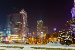 Un paisaje urbano nocturno con edificios iluminados, un parque con bancos y nieve en el suelo.