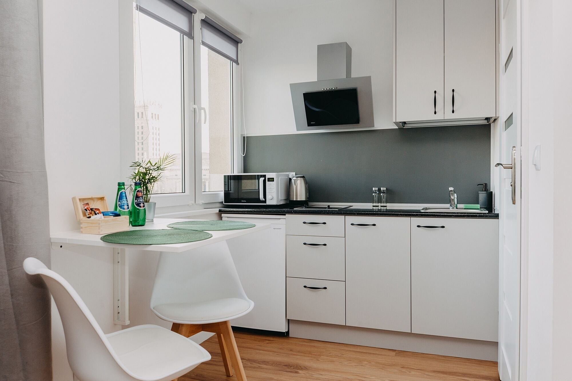 A compact kitchen with white cabinets, a microwave, a kettle, and a small dining area with a white chair and a green mat.