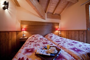 Een gezellige kamer met houten wanden en een hoge plafond, met twee bedden voorzien van beddengoed met een patroon, nachtlampjes en een dienblad met een krant en een kop.