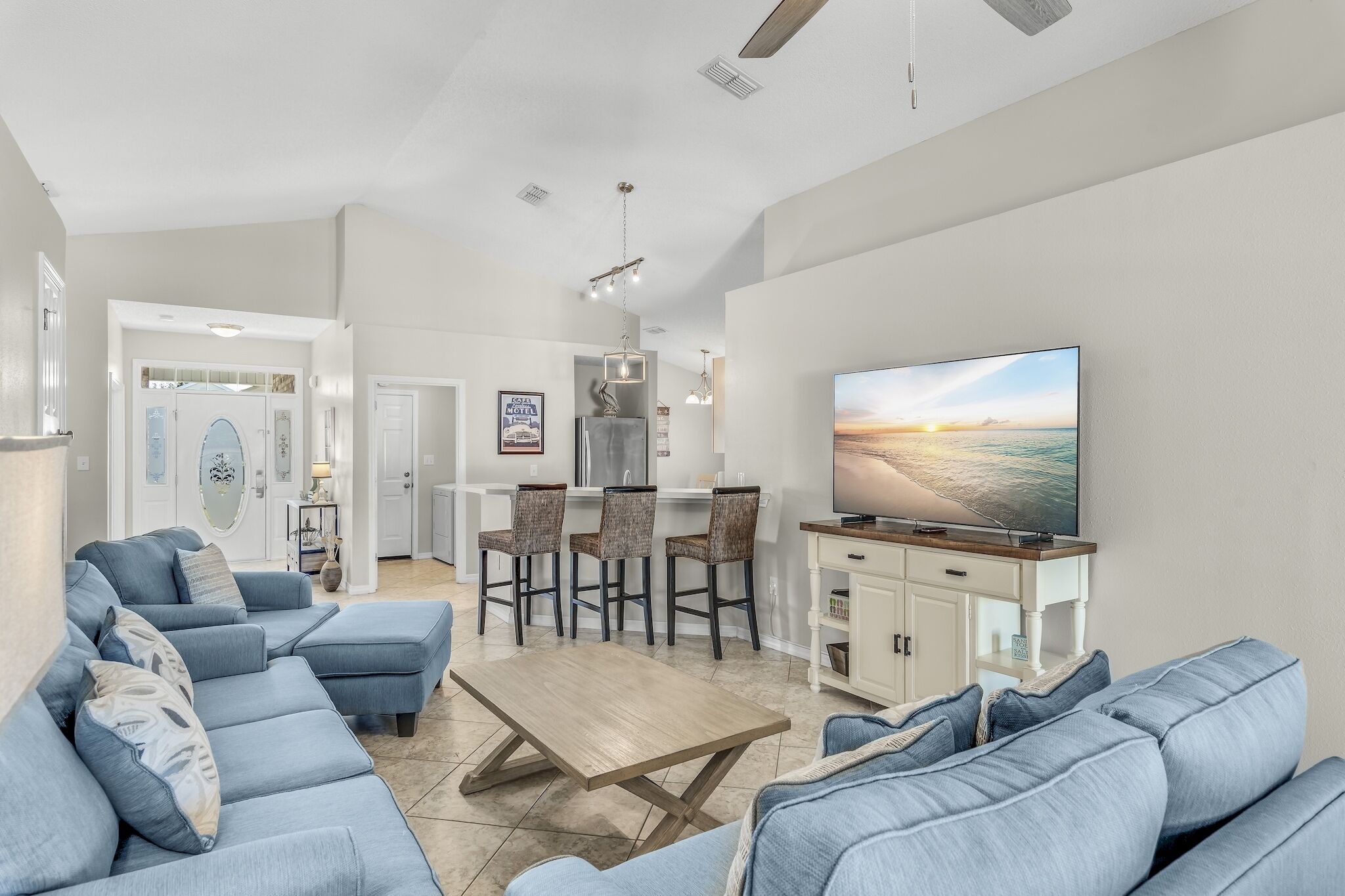 A spacious living room with a large TV, blue sofas, a wooden coffee table, and a kitchen area in the background.