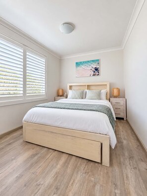 A bedroom with a bed, bedside tables, a painting, and a window with blinds.