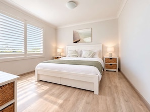 Een slaapkamer met een bed, nachtkastjes, een lamp, een raam met zonwering en een schilderij aan de muur.