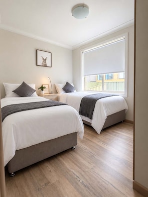A hotel room with two beds, a bedside table with a lamp, a framed picture on the wall, and a large window with a view of a building. - Malua Bay Beach House | Wake Up to the Waves (MALUA BAY)