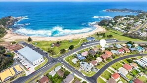 An aerial view of a coastal town with a beach, residential areas, and a large building with a flat roof. - Malua Bay Beach house (MALUA BAY)