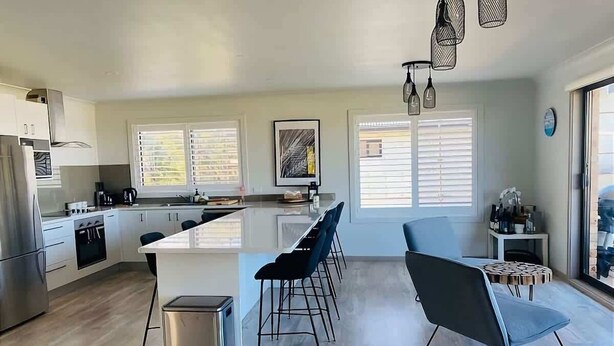 A modern kitchen with a central island, bar stools, a coffee machine, and a dining area with a chair and table. - Malua Bay Beach house (MALUA BAY)