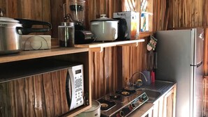 A compact kitchen with wooden walls, a stainless steel refrigerator, a microwave, a stove, and various kitchen appliances on shelves. - Cabin (Provincia de Puntarenas)