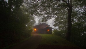 A log cabin with a visible porch and a path leading to it, surrounded by dense trees.