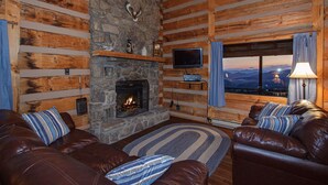 A log cabin interior with a stone fireplace, leather sofas, a television, and a view of the mountains.