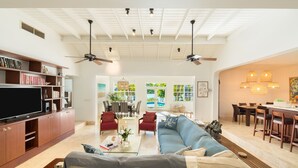A spacious living room with a large sofa, a coffee table, and a TV on a wooden cabinet. There are two ceiling fans, a dining area with a table and chairs, and a kitchen area with a bar. - Water front, with amazing elevated views, pool, flamingoes, private and quiet (Provienciales)