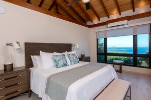 A bedroom with a large bed, wooden ceiling beams, a nightstand, and a view of the ocean through large windows.