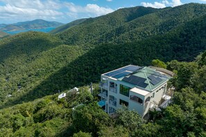 A modern multi-story building with a flat roof, surrounded by dense forest and hills.