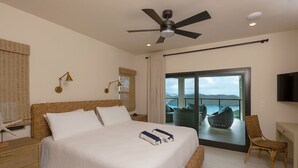 A bedroom with a large bed, a ceiling fan, a TV, and a balcony with a view of the sea.