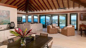 A spacious living area with a high ceiling, large windows offering a view of the sea, and a dining area with a table and chairs.