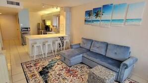 A living room with a grey sofa, a patterned rug, and a kitchen area with white bar stools.