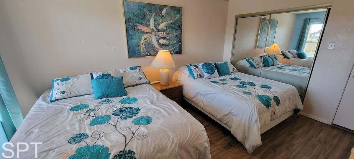 A hotel room with two beds, each with floral-patterned bedding and matching pillows. There is a bedside table with a lamp, a painting on the wall, and a mirror reflecting the room.