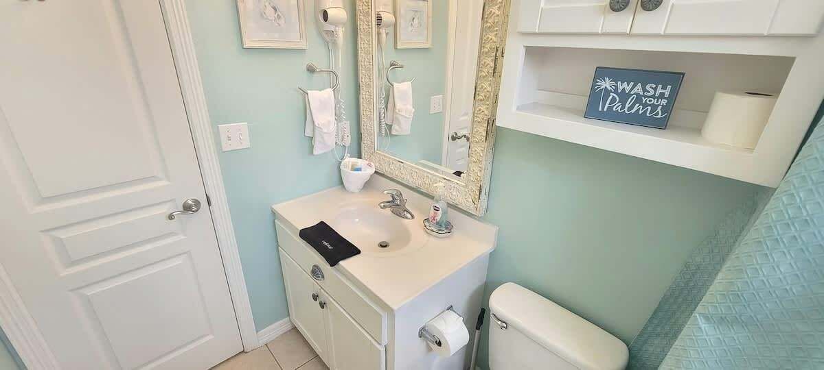 Un baño con lavamanos, espejo, toallero, inodoro y un letrero que dice 'Lava tus manos'.