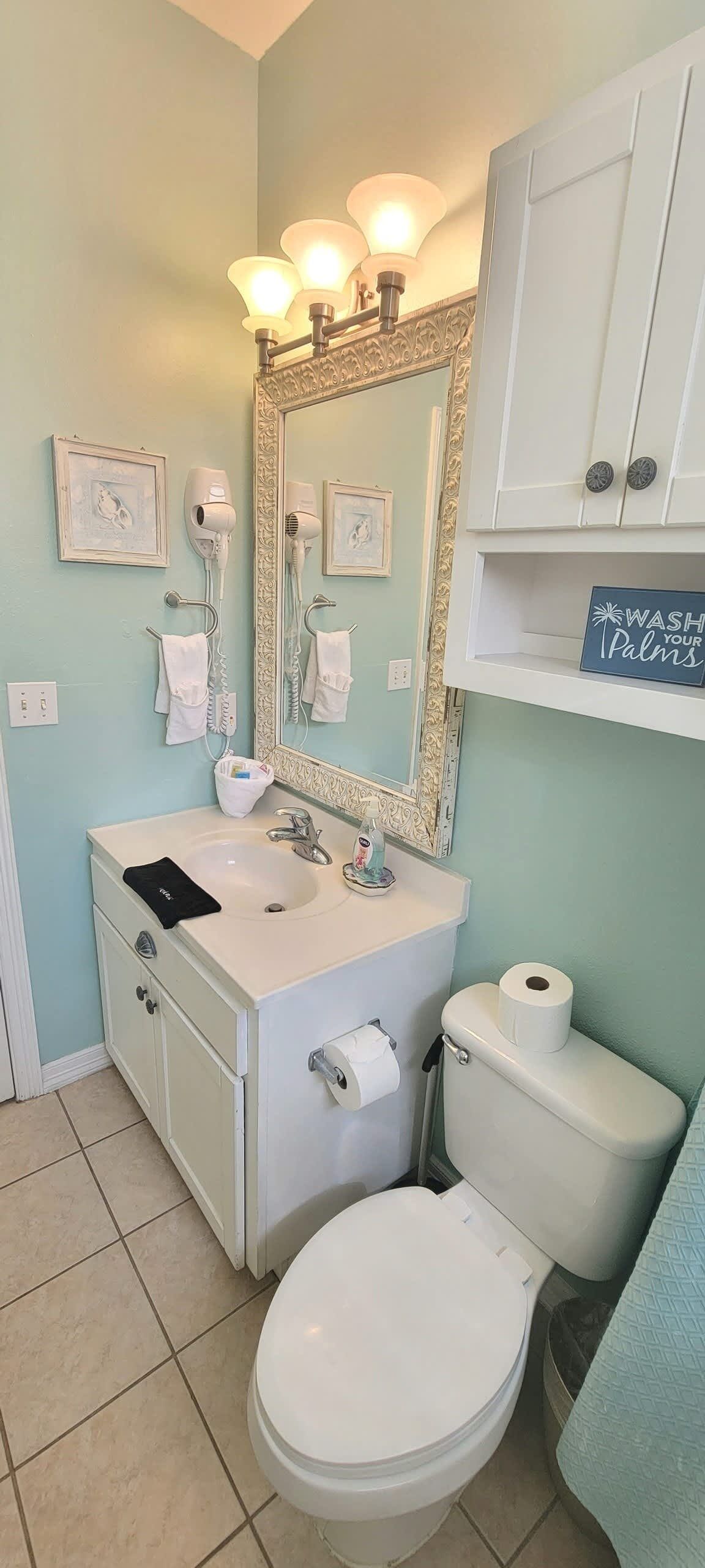 A bathroom with a white sink, a toilet, and a large mirror with lights. There are two hair dryers on the wall, a towel rack, and a sign that says "WASH YOUR PALMS".