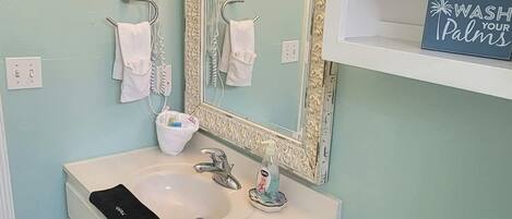 A bathroom with a white sink, a toilet, and a large mirror with lights. There are two hair dryers on the wall, a towel rack, and a sign that says "WASH YOUR PALMS".
