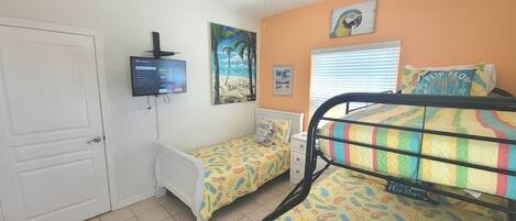 A bedroom with two single beds, a TV, a beach-themed painting, and a window with blinds.