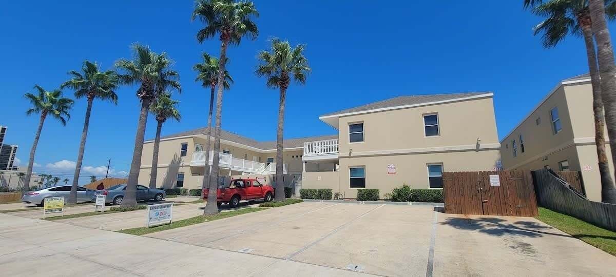 A multi-story building with a parking lot, palm trees, and a red vehicle parked in front.
