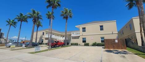 A multi-story building with a parking lot, palm trees, and a red vehicle parked in front.