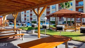 Een terras buiten met houten banken en barbecuegrills, omringd door een zwembad en een meerlaags gebouw met balkons.
