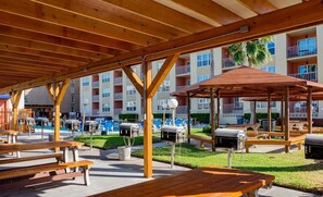 Ein Außenbereich mit einer Terrasse, Holzbanken und Grillstationen, umgeben von einem Pool und einem mehrstöckigen Gebäude mit Balkonen.