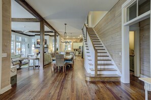 Un interior espacioso con una escalera de madera, un comedor con candelabro, y una cocina visible a través de un vano abierto.