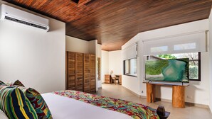 A bedroom with a large bed, an air conditioner, wooden wardrobe, a desk, and a TV mounted on a wooden stand.