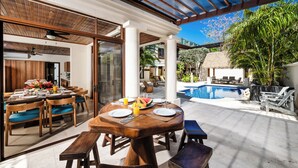 A dining area with a poolside setting, featuring a wooden table and chairs, a thatched-roof structure, and a clear view of the pool.
