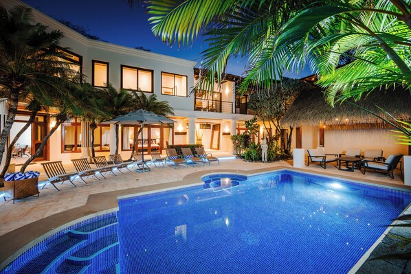 A swimming pool with lounge chairs and umbrellas, surrounded by palm trees and a building with multiple windows.