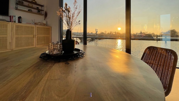 Een eetgedeelte met uitzicht op een rivier en zonsondergang, een vaas met gedroogde bloemen en een houten tafel.