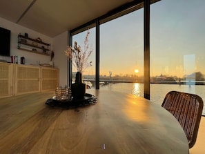 A dining area with a view of a river and sunset, a vase with dried flowers, and a wooden table.