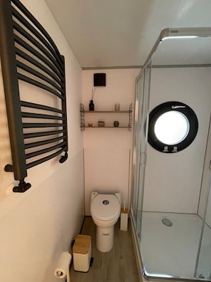 A compact bathroom with a toilet, a shower enclosure, a towel holder, and a small shelf with toiletries.