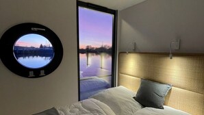 Een slaapkamer met een bed, een ronde spiegel, een raam met uitzicht op een waterpartij, en een hoofdbord.