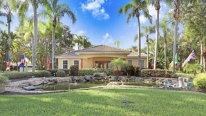 A house with a tiled roof, surrounded by palm trees and a small pond. Flags are visible in the background. - Luxury n cozy 3 bed 2 bath vacation property in a gated community (Kissimmee)