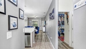 A hallway with framed artwork on the walls, a kitchen area with a dishwasher, and a dining area with blue chairs. - Luxury n cozy 3 bed 2 bath vacation property in a gated community (Kissimmee)
