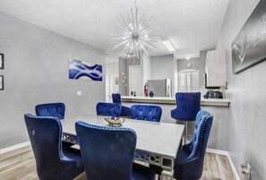 Un comedor moderno con una mesa blanca y sillas tapizadas en azul, una lámpara decorativa en el techo y obras de arte en las paredes.